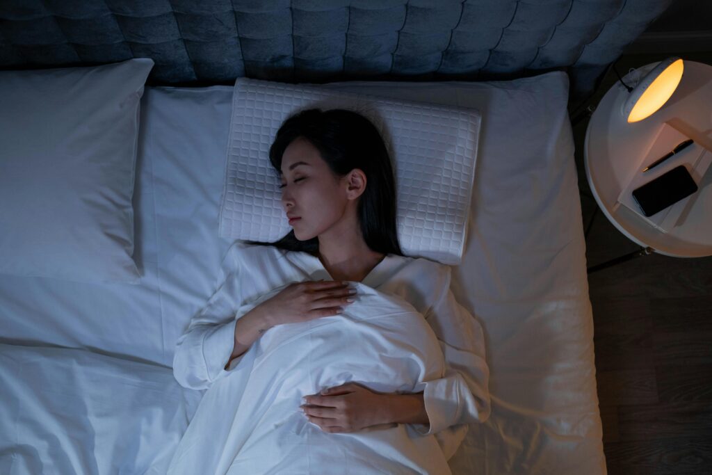 mulher dormindo em uma cama com uma mesa de cabeceira redonda ao lado contendo uma luminária, um caderno, uma caneta e um celular